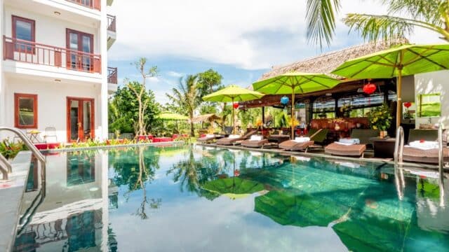 Hoi An resort pool with lounge chairs, green umbrellas, palm trees, and a building with balconies under a bright sky.