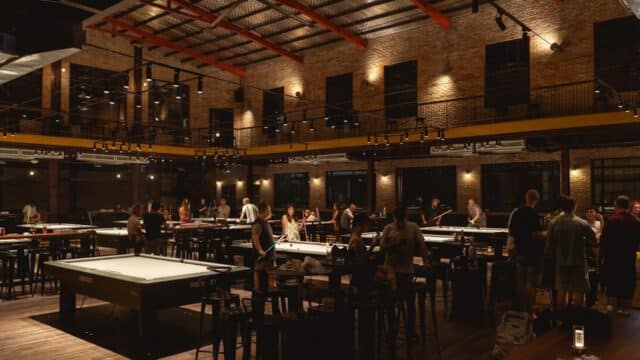 People playing pool in a large, dimly lit hall with exposed brick walls, reminiscent of Hoi An nightlife.