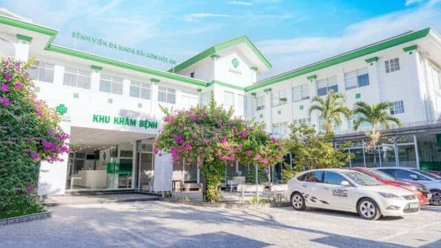 A modern hospital in Hoi An with green accents, blooming bougainvillea, and parked cars on a sunny day.