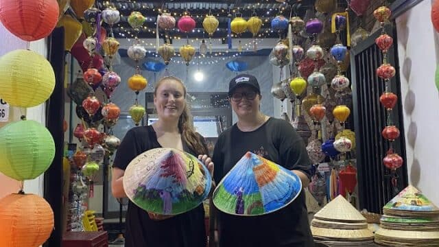 Two people hold painted conical hats, standing under colorful lanterns at a vibrant Hoi An market stall.