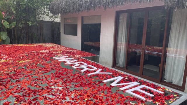 A patio in Hoi An is covered with red rose petals spelling MARRY ME in large white letters near glass doors.