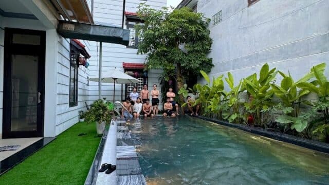 A group of people sit at the edge of a narrow backyard pool between two houses in Hoi An, surrounded by lush plants.