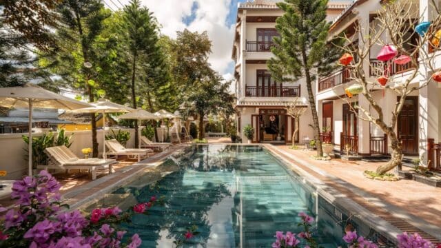 A serene Hoi An hotel pool surrounded by lounge chairs, trees, and colorful lanterns on a sunny day.