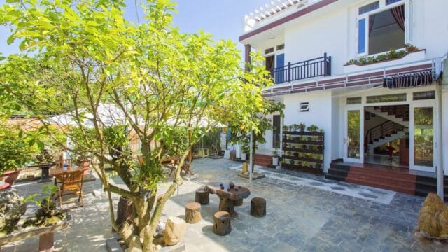 Sunny courtyard with trees, wooden tables and stools, beside a modern white house with large windows, inspired by Hoi An charm.