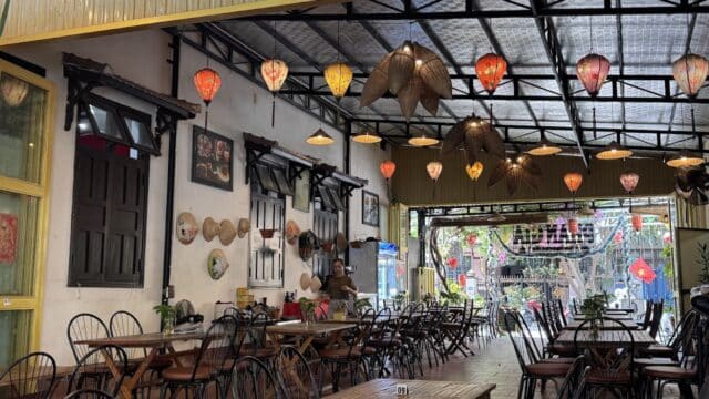 A cozy Hoi An cafe with lanterns and hats hanging from the ceiling, empty wooden tables, chairs, and a charming street view.