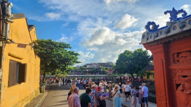 Tourists gather on a sunny street near Hoi An’s river, with yellow buildings and a red decorative structure in the background.