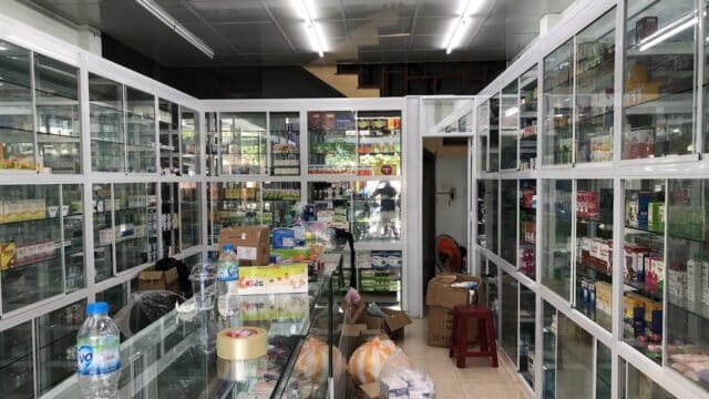 A Hoi An pharmacy with glass shelves of medicine and boxes, scattered items on the counter, and water bottles.