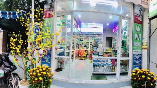 Hoi An glass-front pharmacy adorned with yellow flowers and plants, shelves inside neatly stocked with medicine.