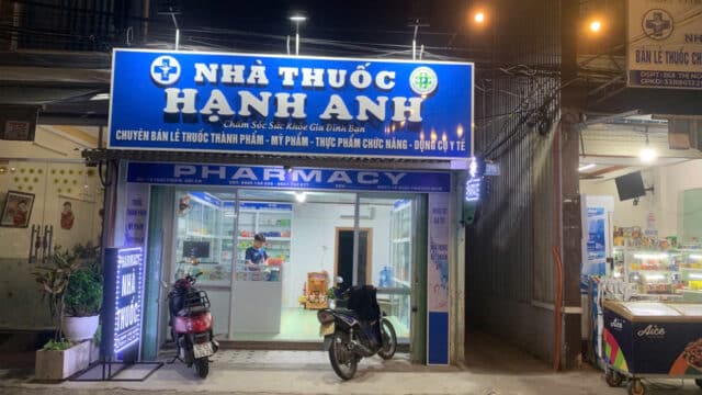 A brightly lit pharmacy named Nhà Thuốc Hạnh Hạnh in Hoi An, with two parked motorbikes in front at night.