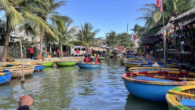 Round boats with people in red shirts float on a river in Hoi An, fringed by palm trees and riverside huts under a blue sky.