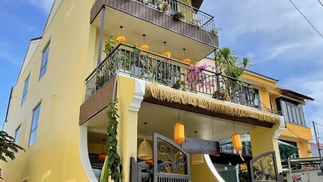 A tall yellow Hoi An house with balconies, lanterns, lush plants, and an open black gate beneath the blue sky.