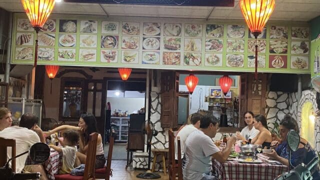 People dining at a cozy Hoi An restaurant with red lanterns, checkered tablecloths, and a menu displayed on the wall above.