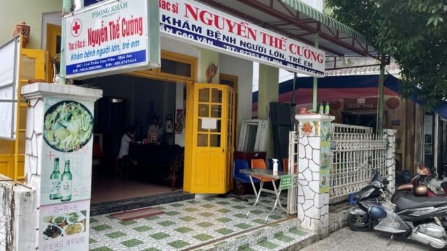 Clinic entrance with yellow doors in Hoi An, signs in Vietnamese, scooters outside, and food photos displayed on the left wall.