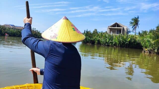 Person in blue shirt and conical hat paddling a boat on a river in Hoi An, with trees and houses in the background.