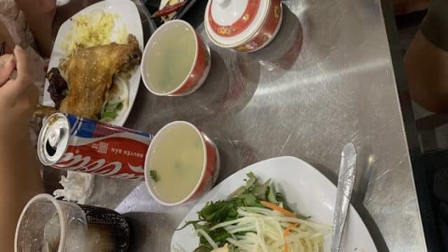 Two plates of rice with chicken, soup bowls, veggies, and Coke cans on a shiny metal table in Hoi An, surrounded by red chairs.