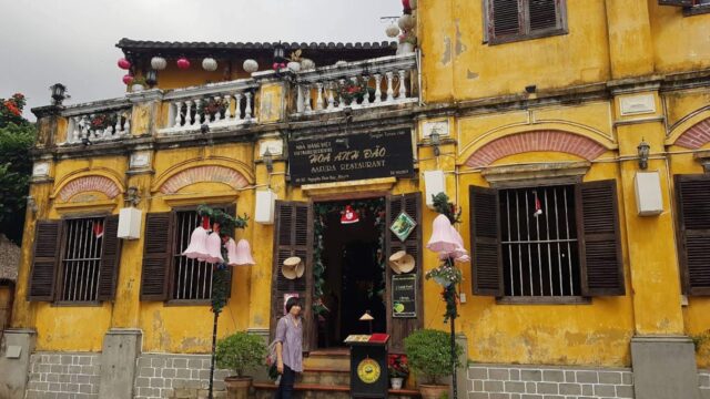 A person walks past a yellow, old-style Hoi An building with wooden shutters and hanging pink lanterns.