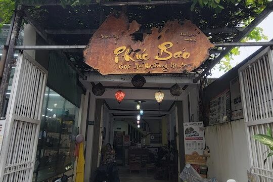 Entrance of a Hoi An cafe or shop with a wooden sign, lush plants above, motorbikes in front, and warm hanging lights inside.