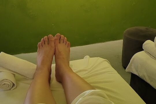 Person lying on a massage bed with feet up, wearing light pants, enjoying relaxation in Hoi An against a green wall background.