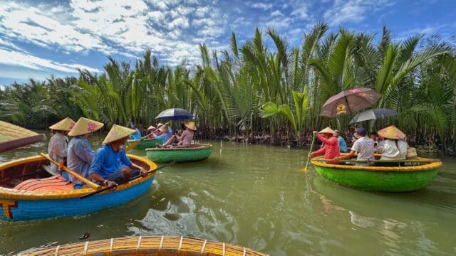 People in round basket boats wearing conical hats paddle the river in Hoi An, lined with palm trees under a blue sky.