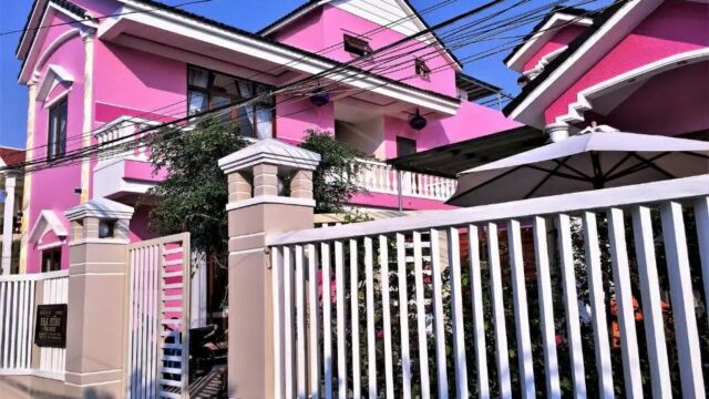 A bright pink two-story house with white trim, a white fence, and power lines overhead in the style of Hoi An on a sunny day.