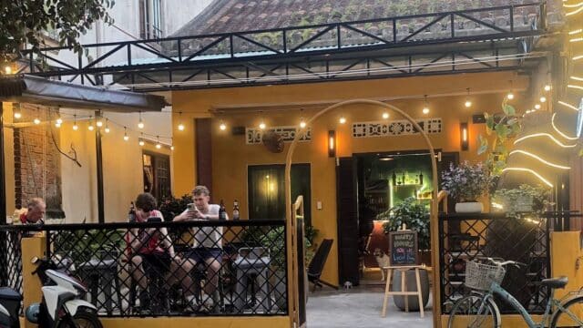 Cozy outdoor cafe in Hoi An with string lights, people at tables, bikes parked, and warm yellow walls glowing at dusk.