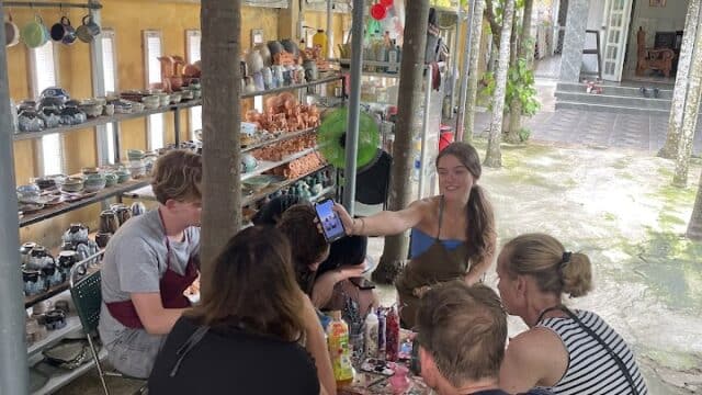 A group of people paint pottery at a table in an outdoor Hoi An studio with shelves of ceramics around them.