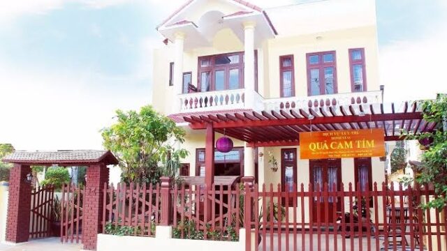 A two-story cream house with red trim, a balcony, red fence, and a yellow QUẢ CAM TÍM sign evokes the charm of Hoi An.