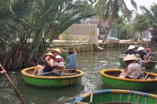 People sit in round basket boats on a river in Hoi An, surrounded by palm trees and greenery, some wearing traditional conical hats.