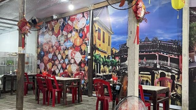Casual eatery with red chairs, wooden tables, Hoi An-inspired wall murals, lanterns, and fans under a metallic ceiling.