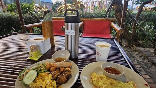 Two plates of food and drinks on a wooden table in a Hoi An outdoor restaurant with garden and lights.