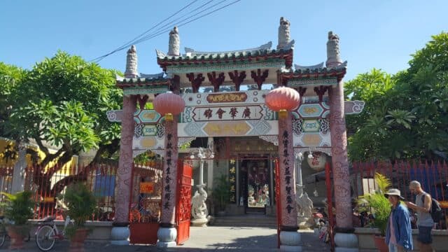 Ornate Chinese temple entrance in Hoi An with red lanterns, intricate carvings, and greenery under a clear blue sky.
