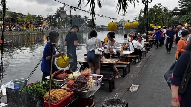 People dining at outdoor riverside food stalls in Hoi An, with skewers, noodles, and vegetables under glowing yellow lanterns.