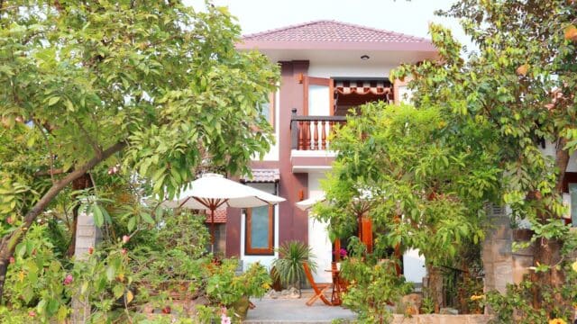 A two-story house with a red roof in Hoi An, surrounded by lush trees, features a patio set and white umbrella out front.