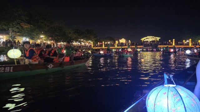Boats with lanterns and people in life jackets float on Hoi An’s river at night, with glowing lights in the background.