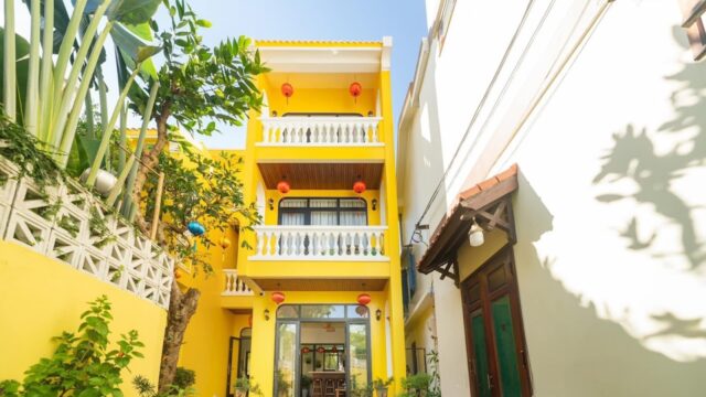 A bright yellow three-story house in Hoi An with balconies, red lanterns, and lush green plants in the courtyard.