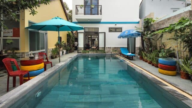 A backyard pool in Hoi An with red chairs, colorful tire stools, lush plants, umbrellas, and a modern two-story house in the background.