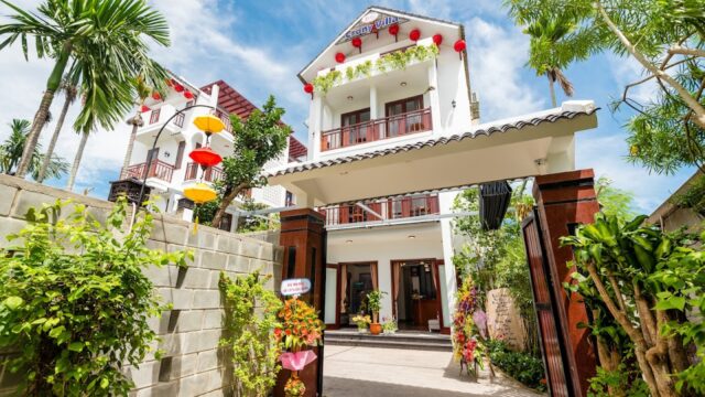 A modern, white three-story Hoi An house with balconies, red lanterns, and a garden entrance under a blue sky.