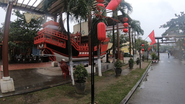A red and black decorative ship in Hoi An, adorned with lanterns and flags, sits beside a wet street lined with palm trees.