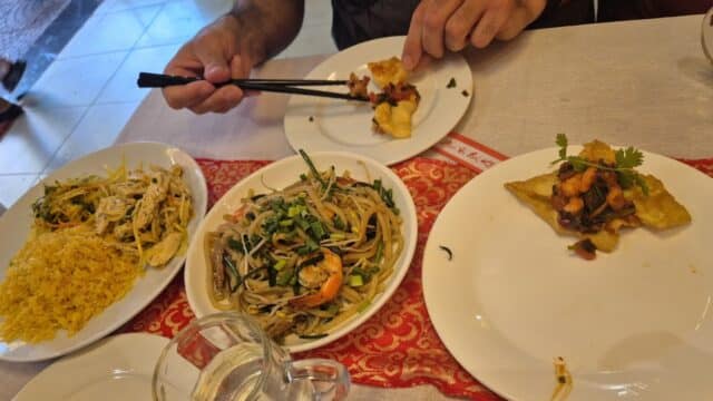 A person uses chopsticks to eat Hoi An Asian dishes, including noodles, fried rice, and appetizers, at a restaurant table.