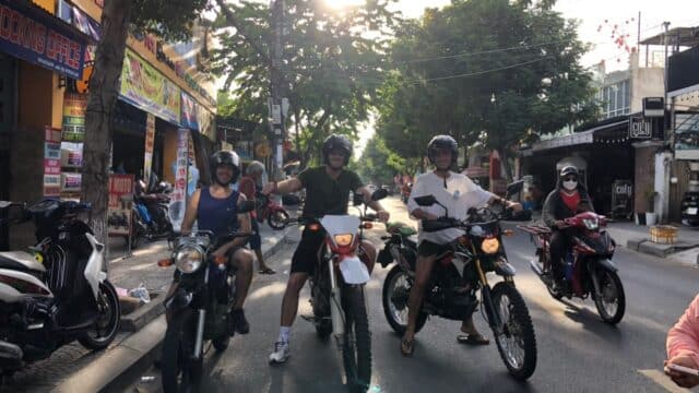 Three people on motorcycles smile and pose in a sunny Hoi An street, with light streaming through trees behind them.