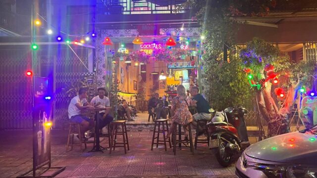 Outdoor bar scene at night in Hoi An with people sitting at tables, colorful string lights, and a lit cocktails sign.