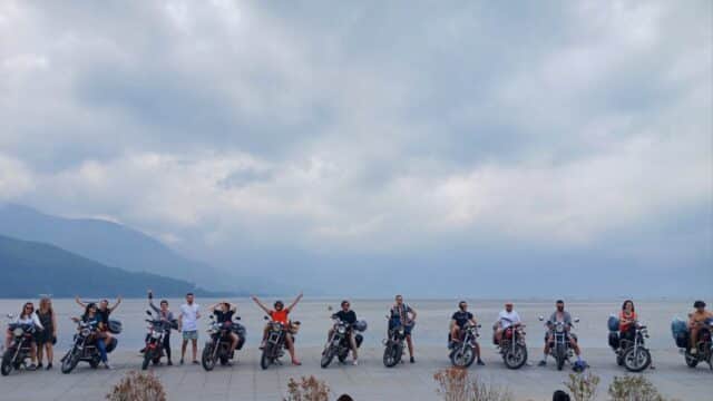 A group of people on motorcycles pose along a seaside promenade in Hoi An, under a cloudy sky, with mountains visible in the background.