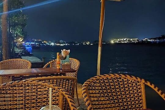 Plate of Vietnamese food and a drink on a wooden table by the river in Hoi An, under an outdoor umbrella at night.