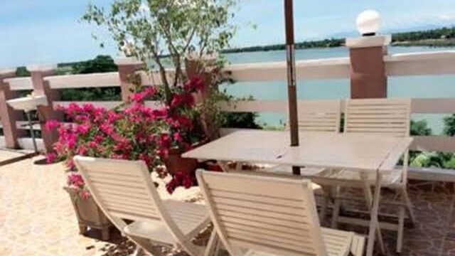 Patio with white chairs and table, pink flowers, and a river view under Hoi An’s sunny blue sky.
