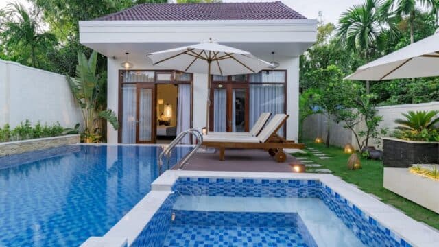 Modern Hoi An villa with private pool, jacuzzi, wooden deck, lounge chairs, and lush garden enclosed by trees and white walls.