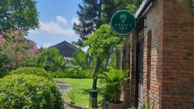 A brick building with a Rosara Spa sign in Hoi An is surrounded by lush greenery and flowering plants under a sunny sky.