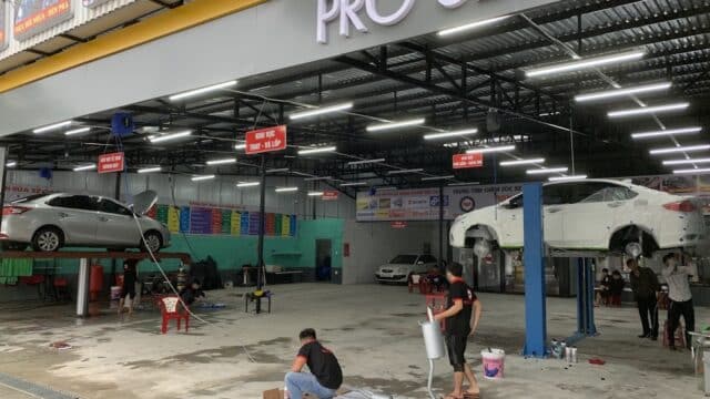 Car service garage in Hoi An with two cars lifted for repairs, workers fixing and painting, and equipment scattered around the open workshop.