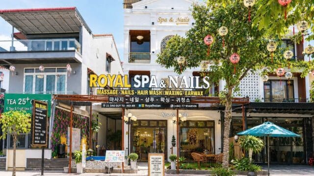 Street view of Royal Spa & Nails shop in Hoi An with trees, lanterns, and outdoor seating in front.
