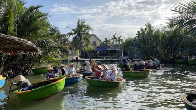 People in conical hats row round basket boats on a river near Hoi An, framed by lush green palms and a partly cloudy sky.