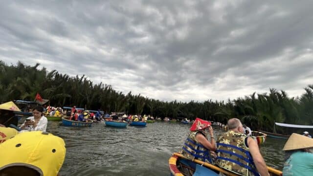 Groups of people in round boats on a river in Hoi An, surrounded by lush greenery under a cloudy sky.